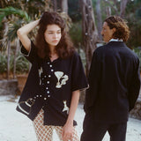 Woman standing outdoors with one hand in her hair, wearing a black embroidered shirt and cream open-knit pants. A man in a black suit stands behind her, facing away. The setting is tropical, with palm trees, string lights, and sandy ground.