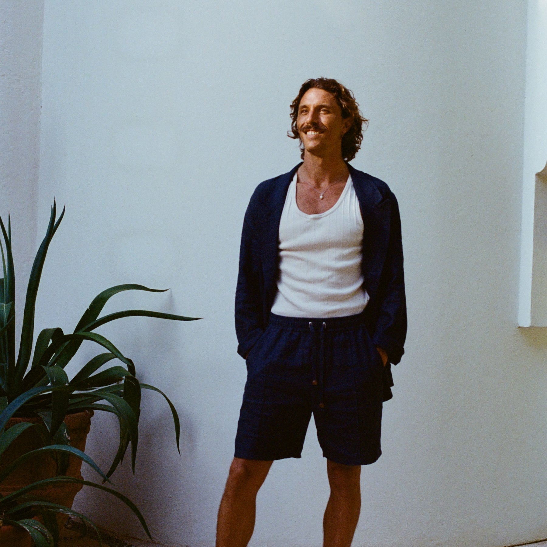 Man standing against a white wall wearing navy linen shorts with a drawstring, a matching navy jacket, a white tank top, and black sandals.