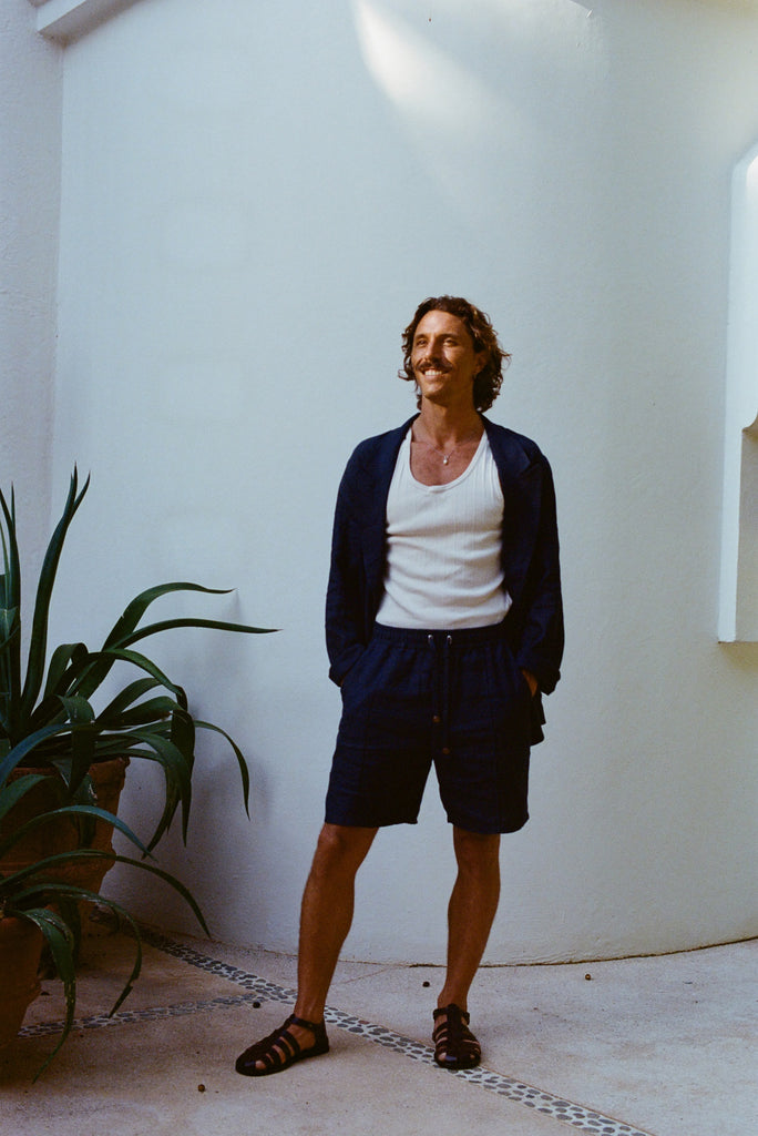 Man standing against a white wall wearing navy linen shorts with a drawstring, a matching navy jacket, a white tank top, and black sandals.