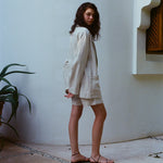 Brunette woman standing sideways, wearing the sand linen Easy Going Jacket with the sand linen Easy Going Shorts and black sandals. She is looking back to the camera. 