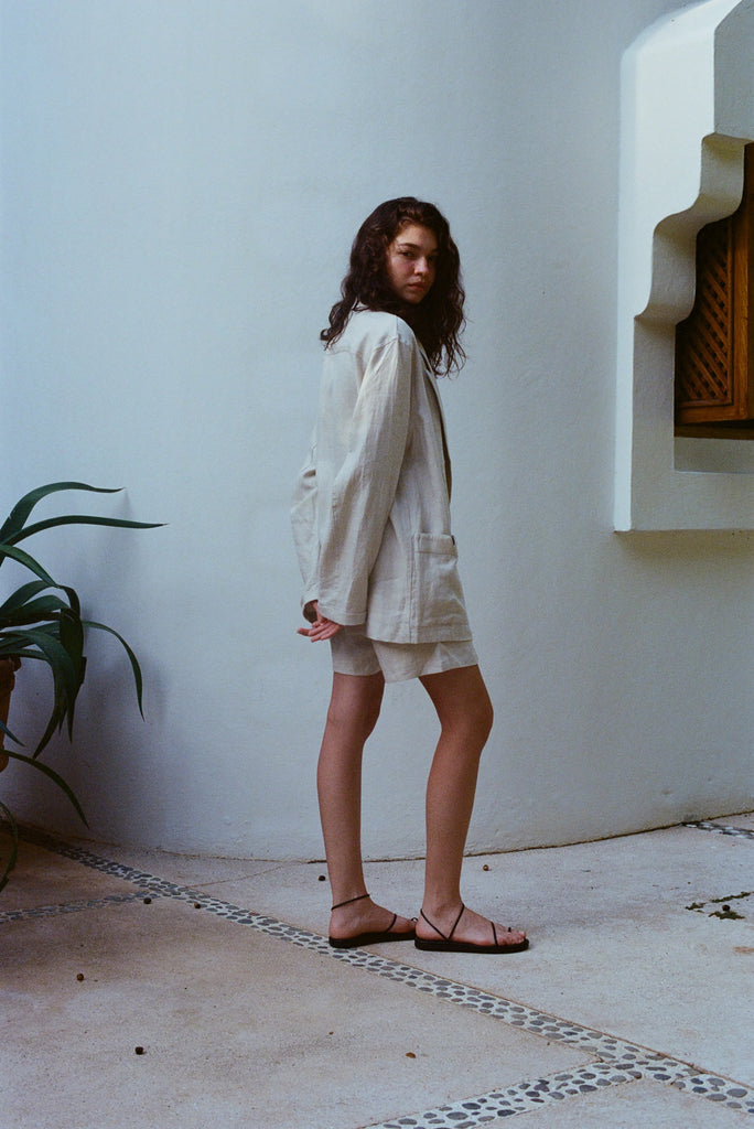 Brunette woman standing sideways, wearing the sand linen Easy Going Jacket with the sand linen Easy Going Shorts and black sandals. She is looking back to the camera. 