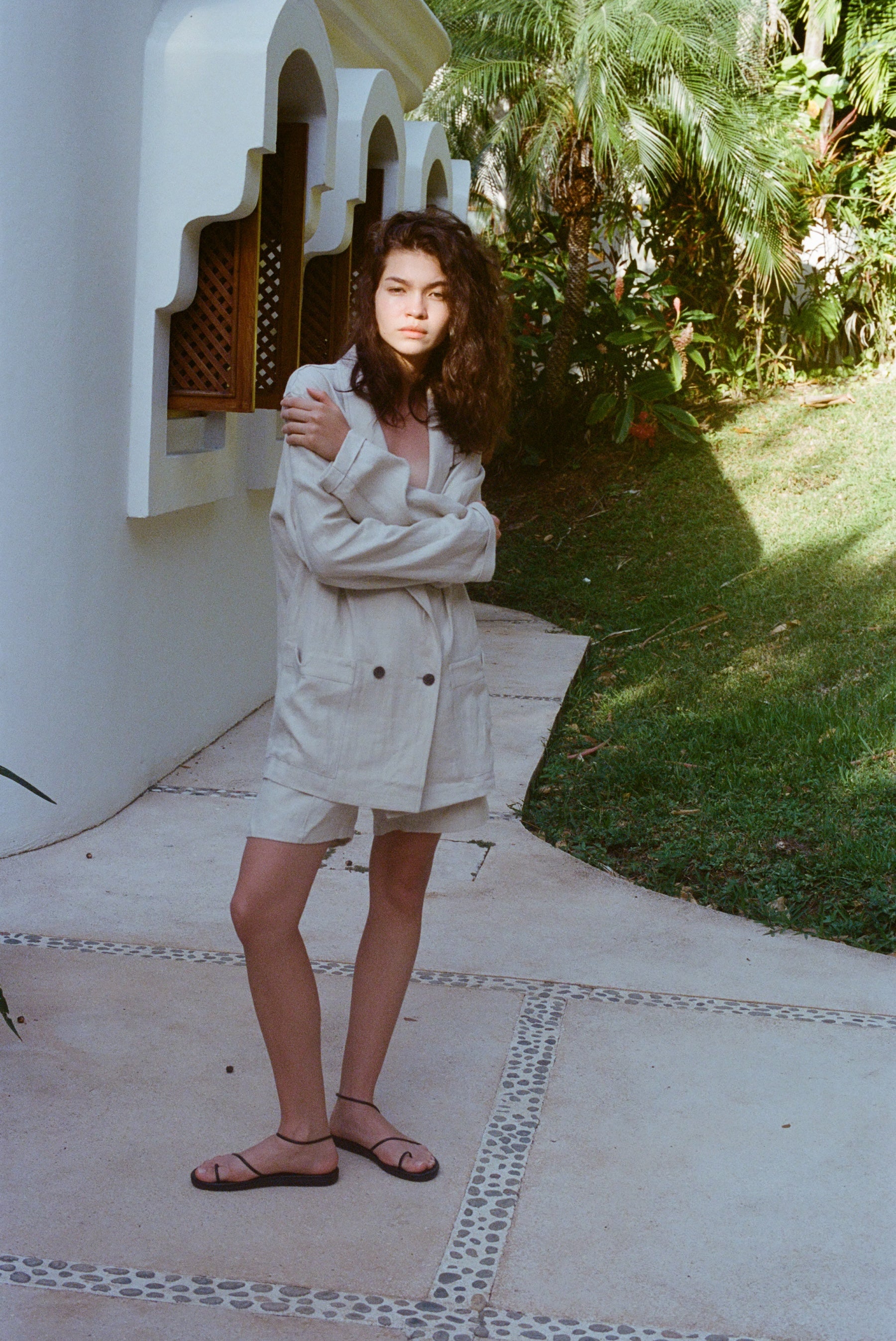 Brunette woman standing in the full Easy Going Linen Suit in Sand with black sandals. She has her arms wrapped around herself.