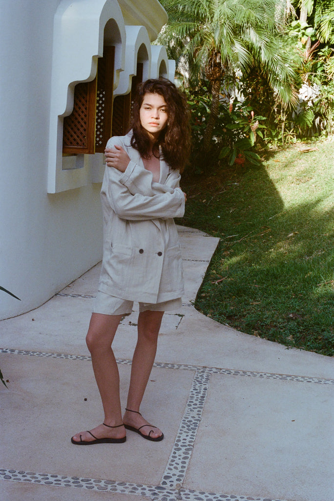 A woman stands wearing the Sand Linen Jacket oversized, with matching Sand linen shorts and black sandals. 
