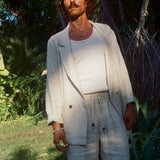 A man stands in partial sunlight wearing a cream-colored linen blazer, matching drawstring shorts, and a white tank top. He has wavy hair and a mustache, and is surrounded by tropical greenery.