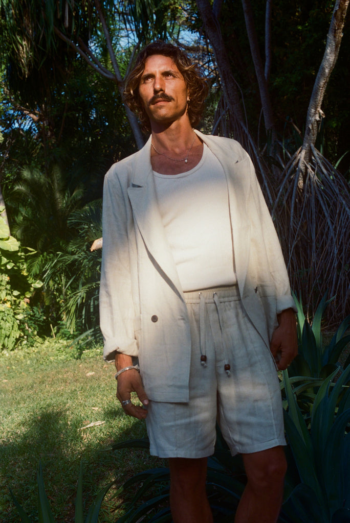 A man stands in partial sunlight wearing a cream-colored linen blazer, matching drawstring shorts, and a white tank top. He has wavy hair and a mustache, and is surrounded by tropical greenery.