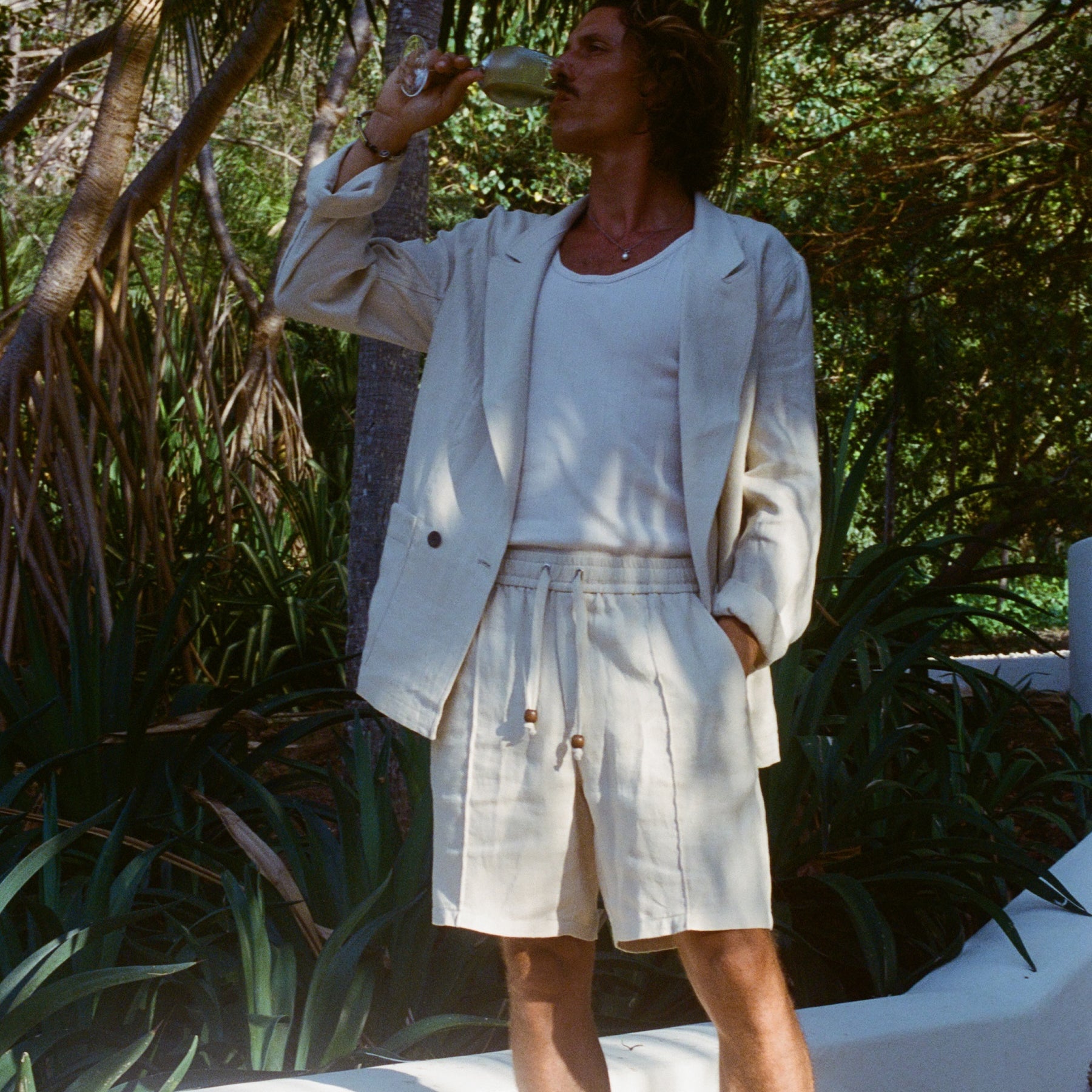 Brunette man sipping on a glass of white wine while wearing a sand linen Easy Going Suit Jacket with sand linen matching shorts, and a while tank top underneath. 