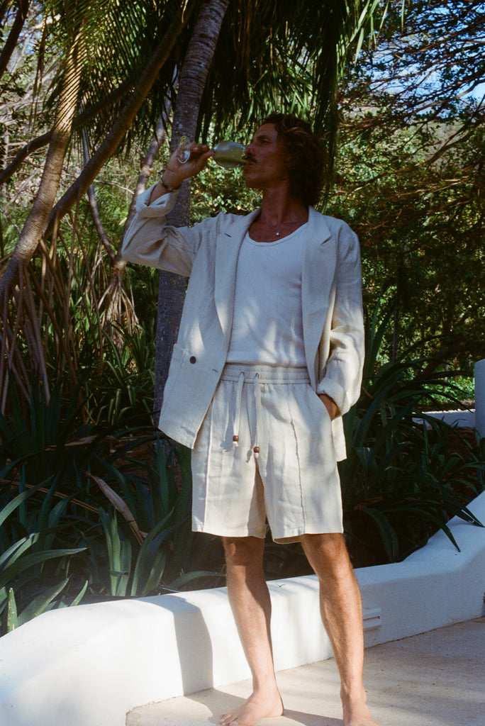 Brunette man sipping on a glass of white wine while wearing a sand linen Easy Going Suit Jacket with sand linen matching shorts, and a while tank top underneath. 