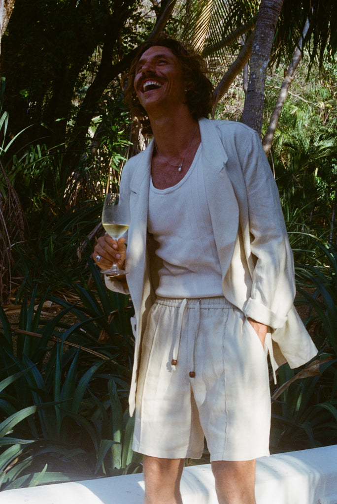 Brunette man with a glass of wine while wearing a sand linen Easy Going Suit Jacket with sand linen matching shorts, and a while tank top underneath. 
