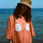 Woman wearing orange sand dollar bikini button down shirt with straw hat, standing in front of the ocean.