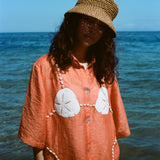 Woman wearing orange sand dollar bikini button down shirt with straw hat, standing in front of the ocean.