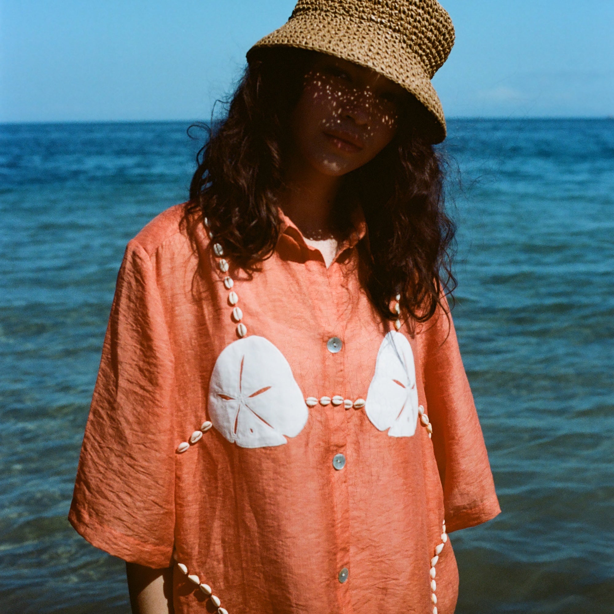 Woman wearing orange sand dollar bikini button down shirt with straw hat, standing in front of the ocean.