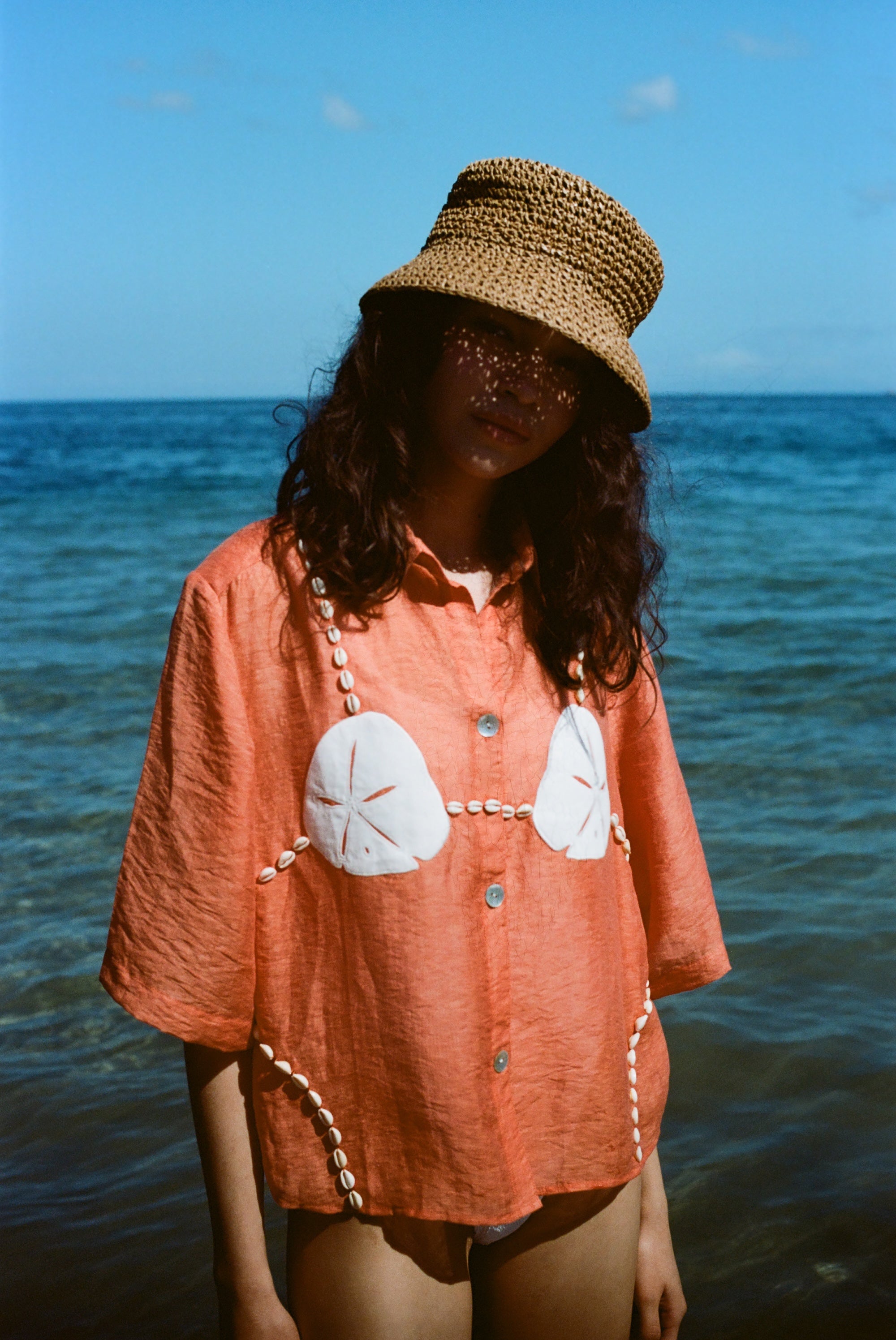 Woman wearing orange sand dollar bikini button down shirt with straw hat, standing in front of the ocean.