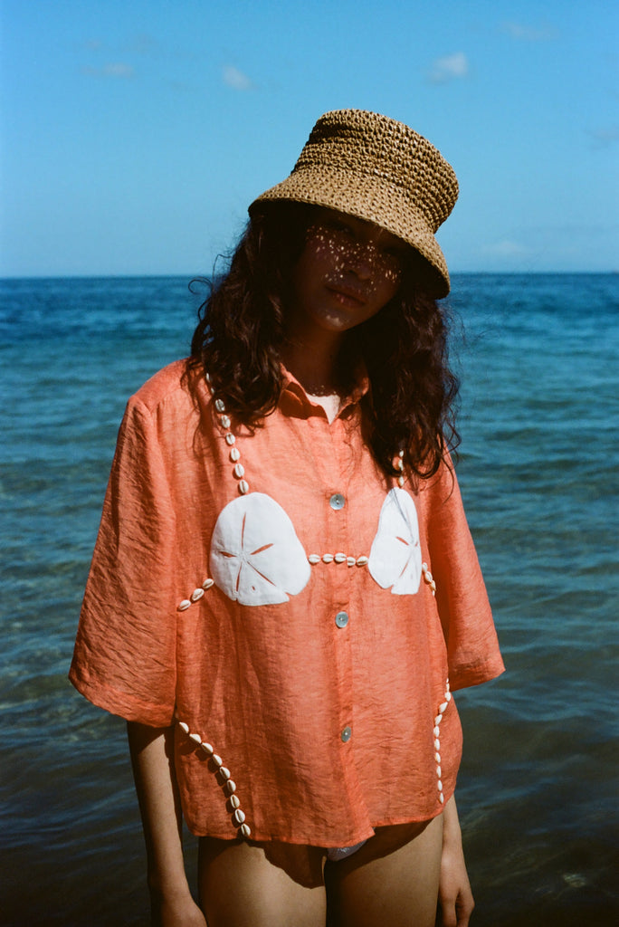 Woman wearing orange sand dollar bikini button down shirt with straw hat, standing in front of the ocean.
