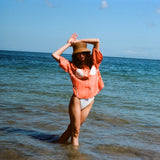 Woman posed with her hands above her head, wearing the sand dollar bikini shirt with a straw hat. She is standing ankle deep in the ocean.
