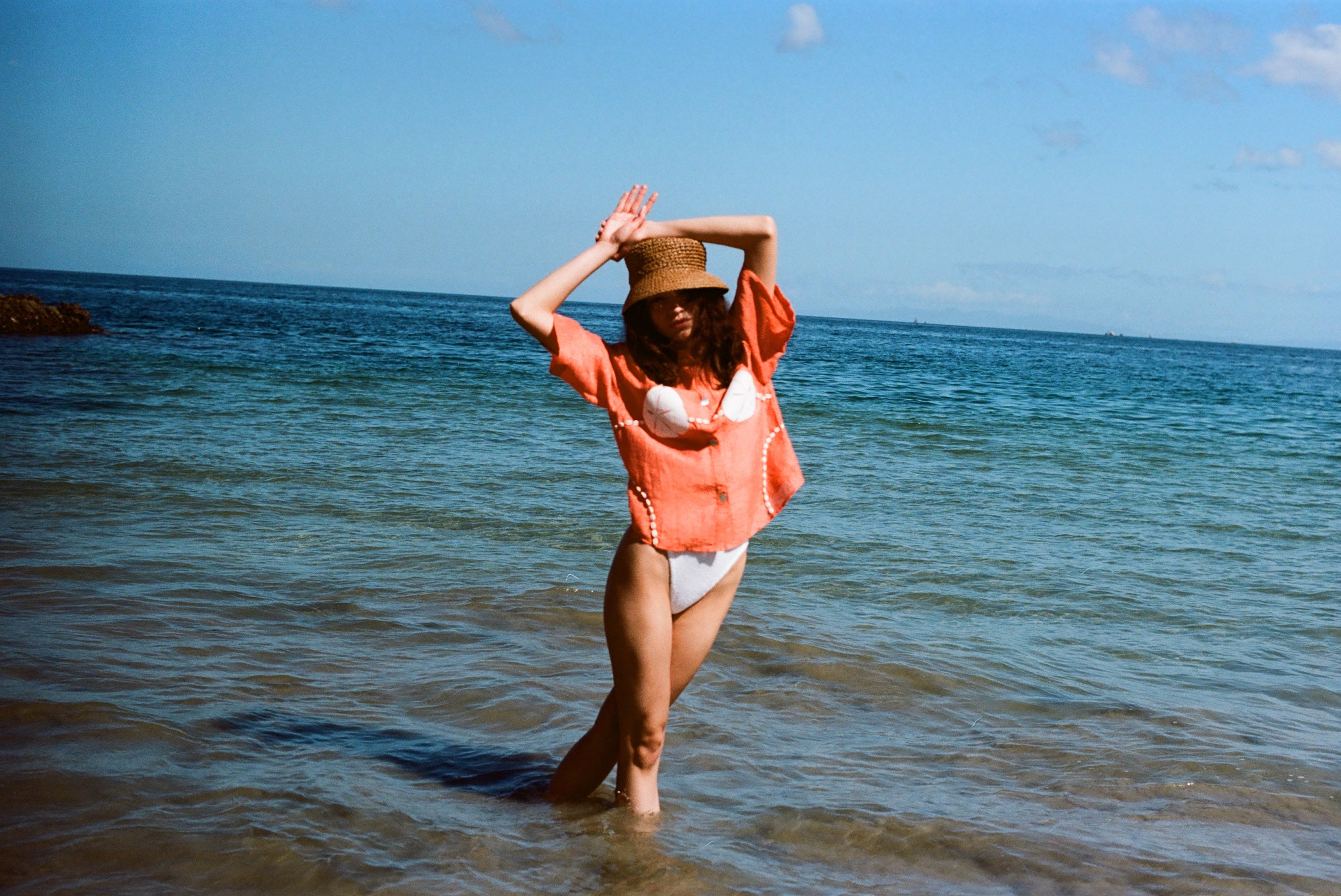Woman posed with her hands above her head, wearing the sand dollar bikini shirt with a straw hat. She is standing ankle deep in the ocean.