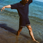Man wearing navy 'TomPolo' and navy swim trunks, dancing ankle deep in the ocean.