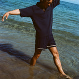 Man wearing navy 'TomPolo' and navy swim trunks, dancing ankle deep in the ocean.