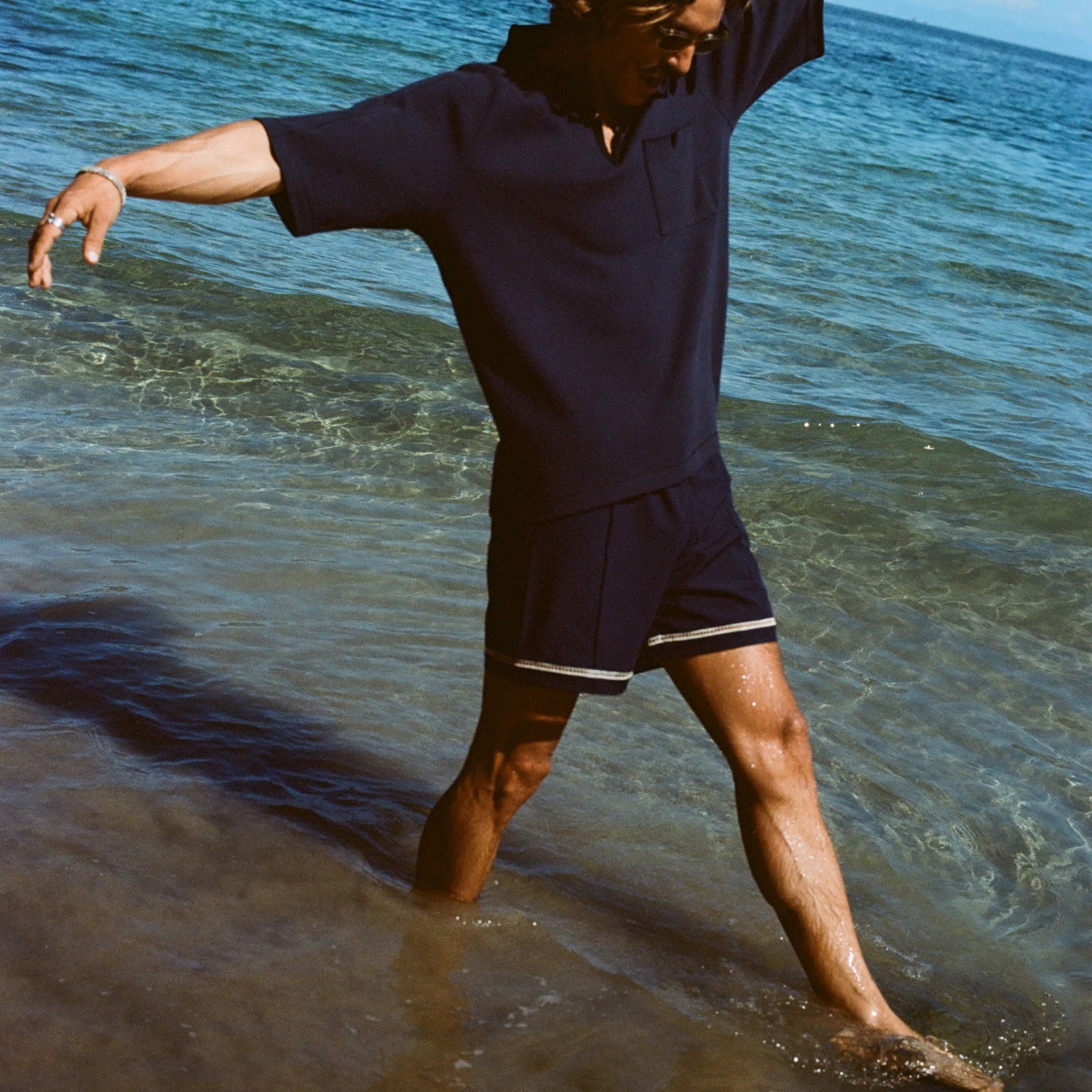 Man wearing navy 'TomPolo' and navy swim trunks, dancing ankle deep in the ocean.