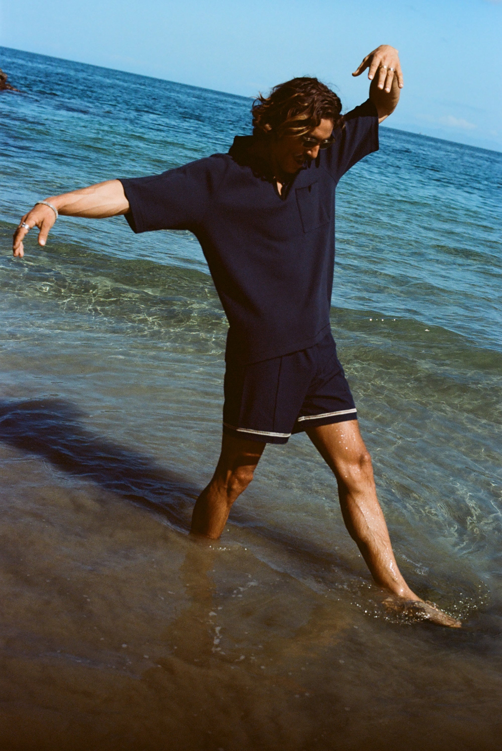 Man wearing navy 'TomPolo' and navy swim trunks, dancing ankle deep in the ocean.
