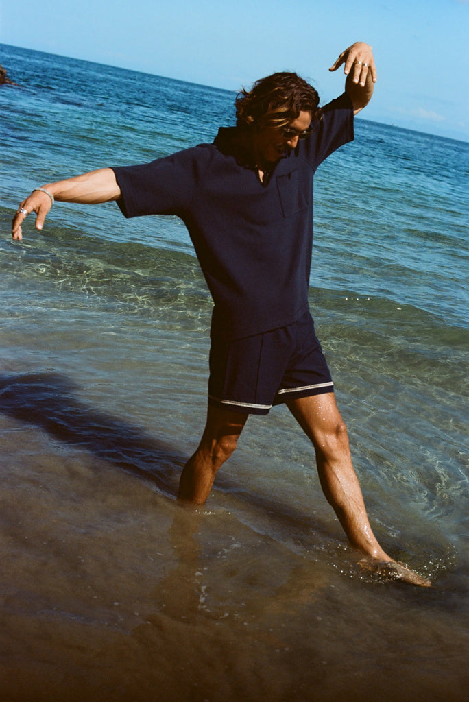 Man wearing navy 'TomPolo' and navy swim trunks, dancing ankle deep in the ocean.