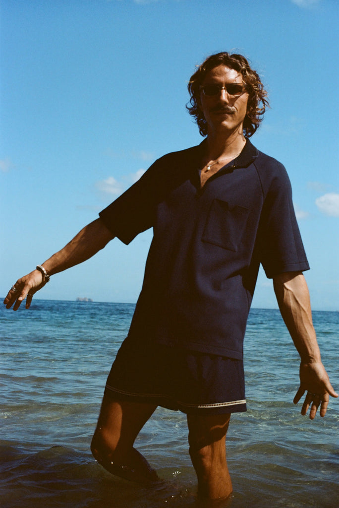 Man posed knee deep in the ocean wearing the navy 'TomPolo' with our navy Cabrisa swim trunks.