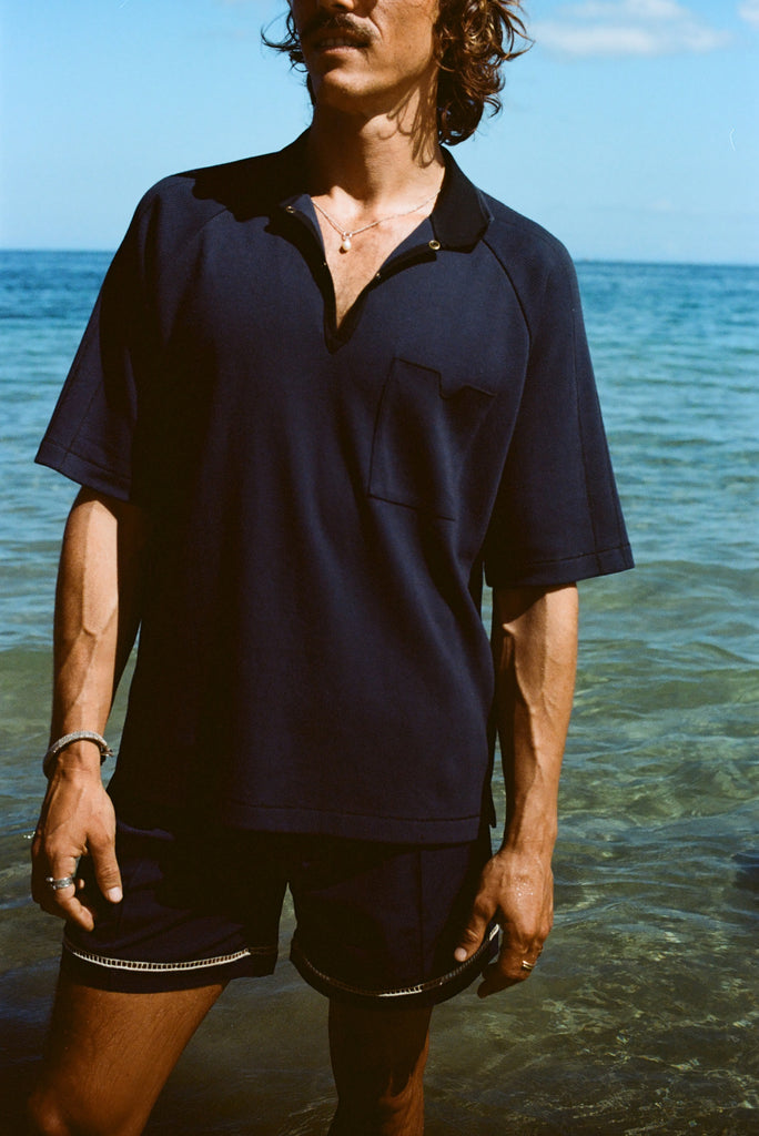 Cropped image of man wearing navy 'TomPolo' with our navy Cabrisa shorts, standing ankle deep in the ocean.