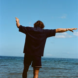 Image from behind of man wearing navy 'TomPolo' and navy swim trunks, ankle deep in the ocean trying to keep balance. 
