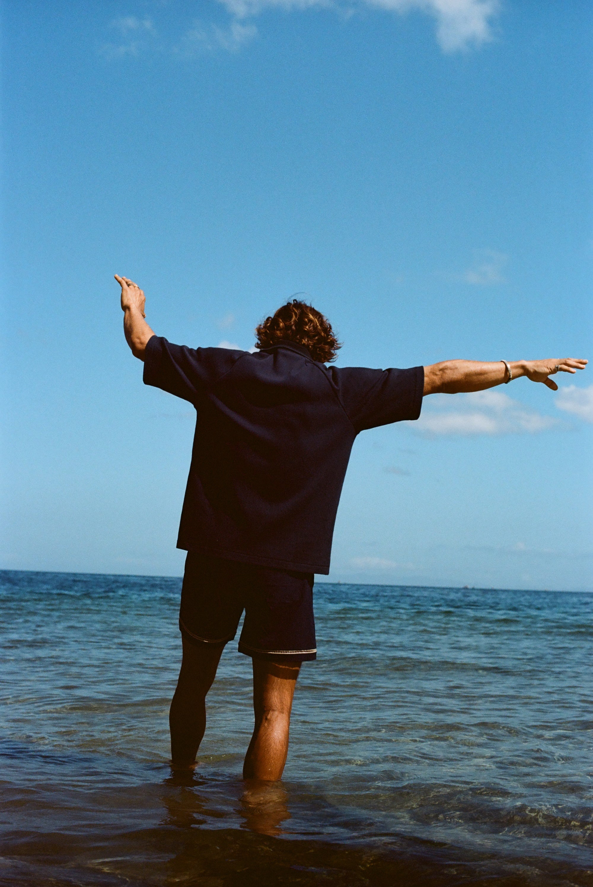 Image from behind of man wearing navy 'TomPolo' and navy swim trunks, ankle deep in the ocean trying to keep balance. 