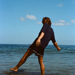 Image from behind of man wearing navy 'TomPolo' and navy swim trunks, ankle deep in the ocean trying to keep balance. 