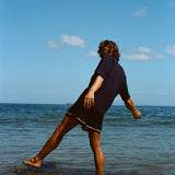 Image from behind of man wearing navy 'TomPolo' and navy swim trunks, ankle deep in the ocean trying to keep balance. 
