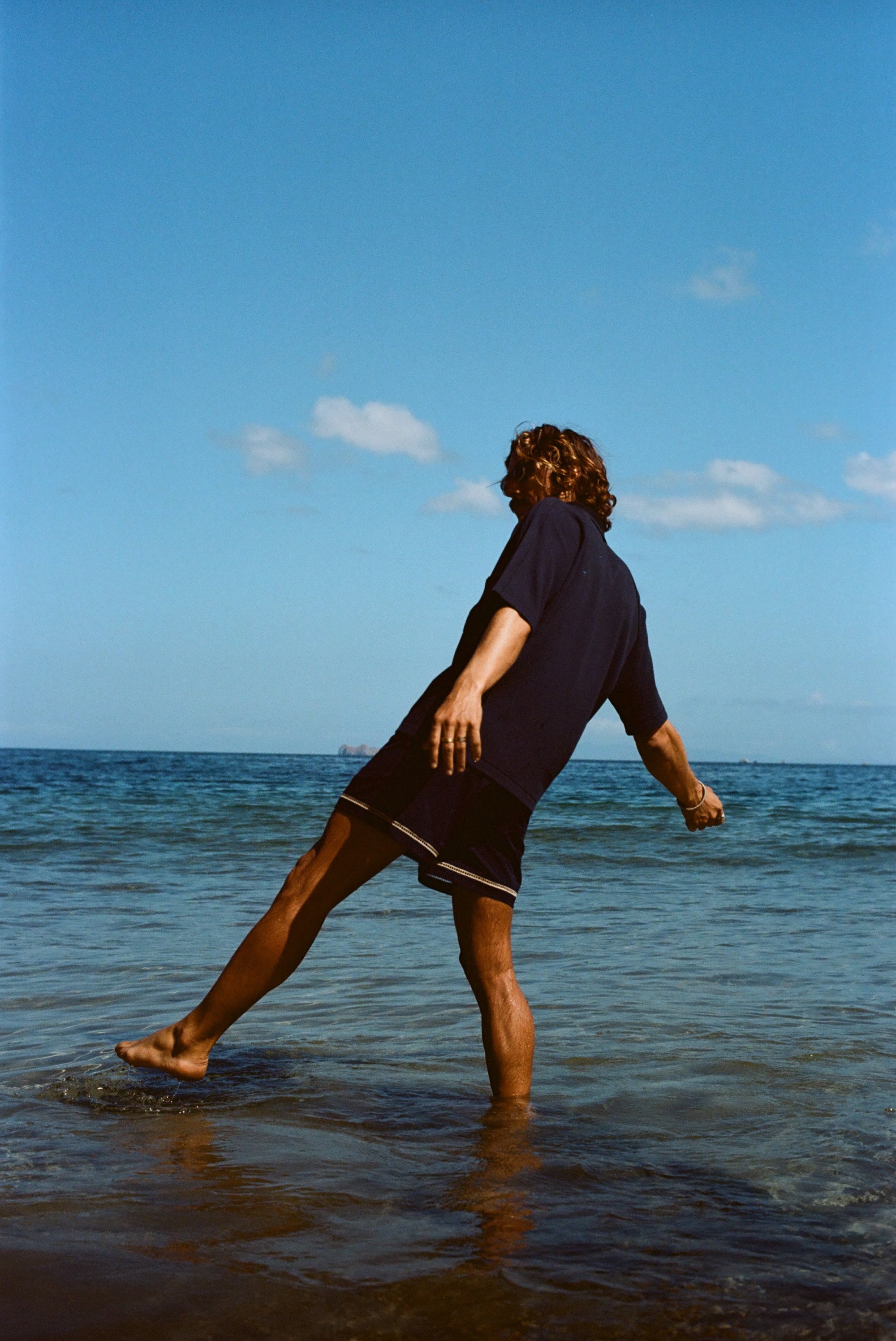 Image from behind of man wearing navy 'TomPolo' and navy swim trunks, ankle deep in the ocean trying to keep balance. 