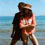 Two people stand on a sunny beach. The woman in front wears the coral button-up shirt with embroidered sand dollars and shell details, a straw hat, and white bikini bottoms. She stands with her arms crossed. Behind her, a man in sunglasses and a dark beach set stands partially in shadow, looking off to the side.