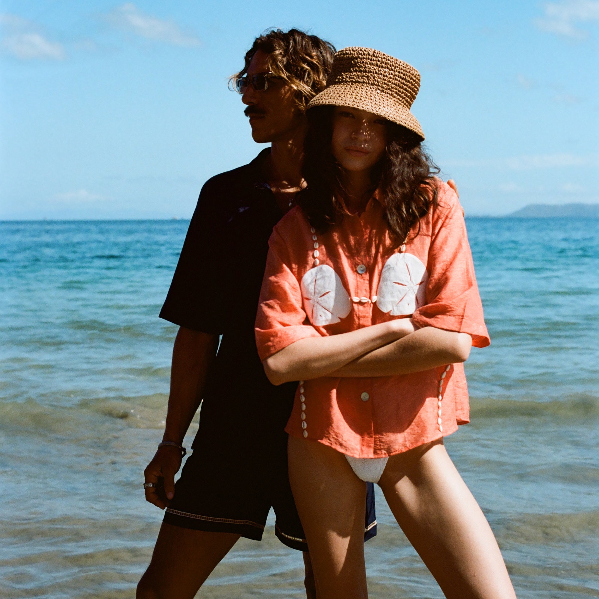 Two people stand on a sunny beach. The woman in front wears the coral button-up shirt with embroidered sand dollars and shell details, a straw hat, and white bikini bottoms. She stands with her arms crossed. Behind her, a man in sunglasses and a dark beach set stands partially in shadow, looking off to the side.