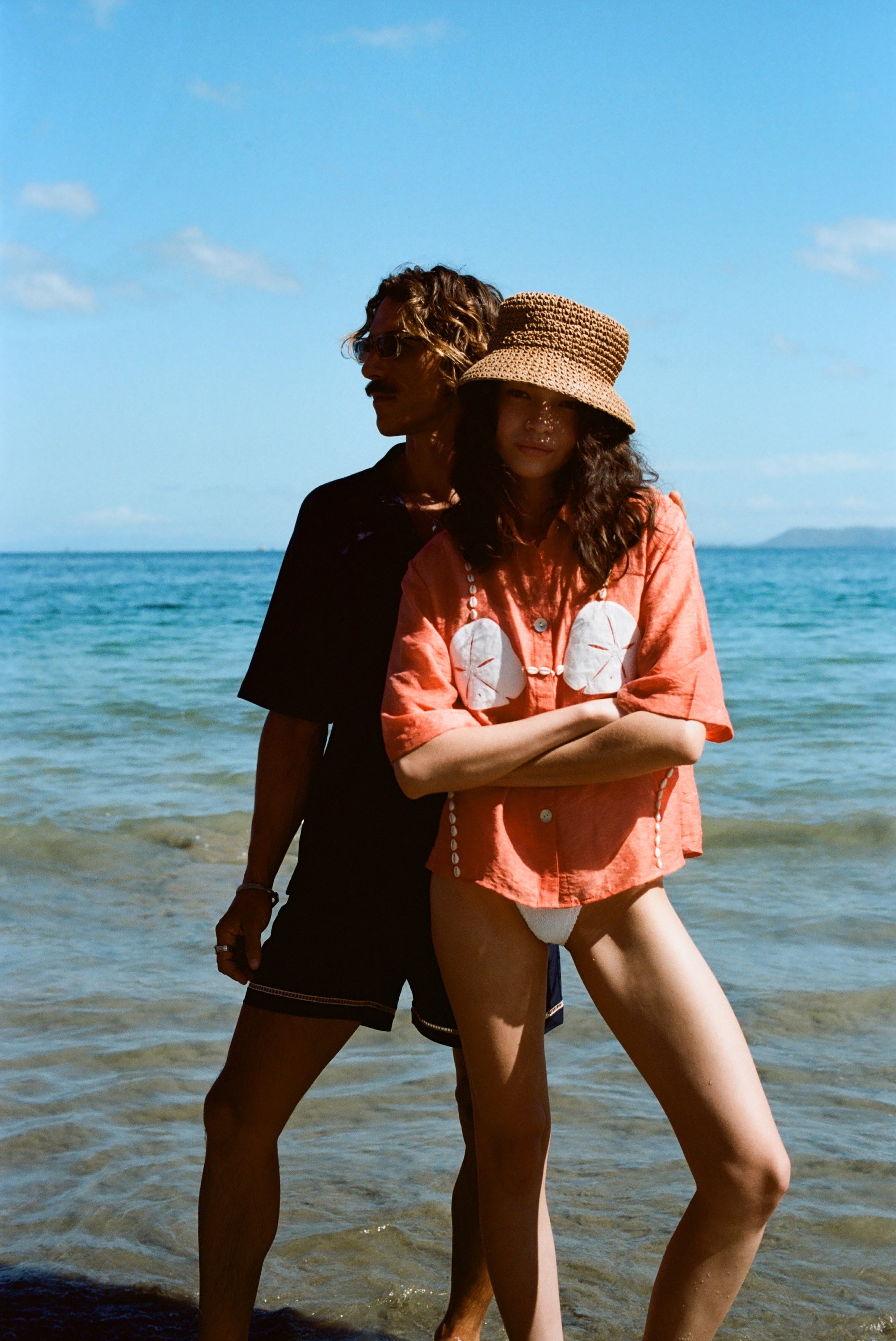 Two people stand on a sunny beach. The woman in front wears the coral button-up shirt with embroidered sand dollars and shell details, a straw hat, and white bikini bottoms. She stands with her arms crossed. Behind her, a man in sunglasses and a dark beach set stands partially in shadow, looking off to the side.