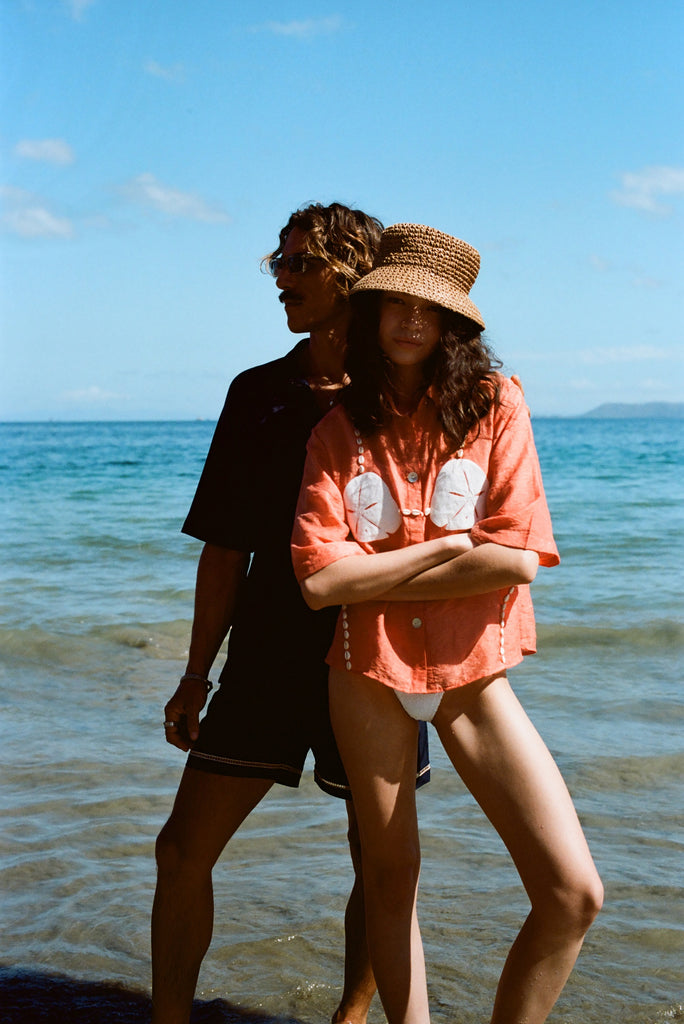 Two people stand on a sunny beach. The woman in front wears the coral button-up shirt with embroidered sand dollars and shell details, a straw hat, and white bikini bottoms. She stands with her arms crossed. Behind her, a man in sunglasses and a dark beach set stands partially in shadow, looking off to the side.