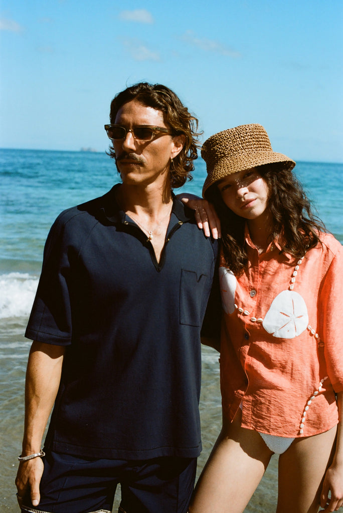 Man and woman posing on the beach wearing the navy mesh 'TomPolo' and our Starfish Strut Shirt in orange. 