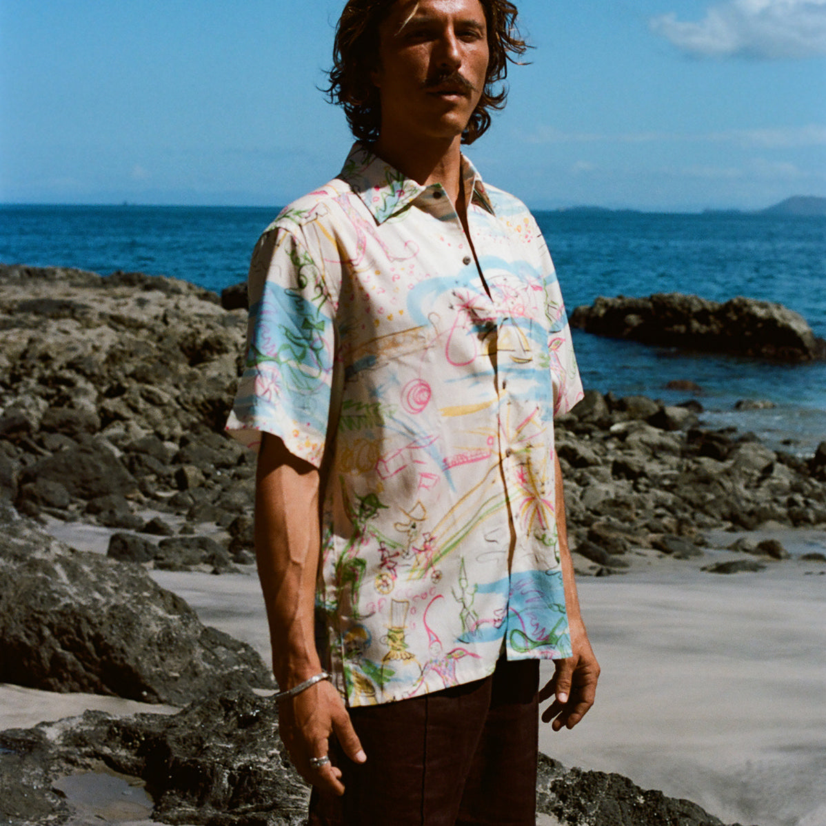 Cream-colored short-sleeve button-down shirt featuring an abstract print in pink, blue, yellow, and green, worn with dark brown shorts.