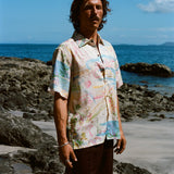 Cream-colored short-sleeve button-down shirt featuring an abstract print in pink, blue, yellow, and green, worn with dark brown shorts.