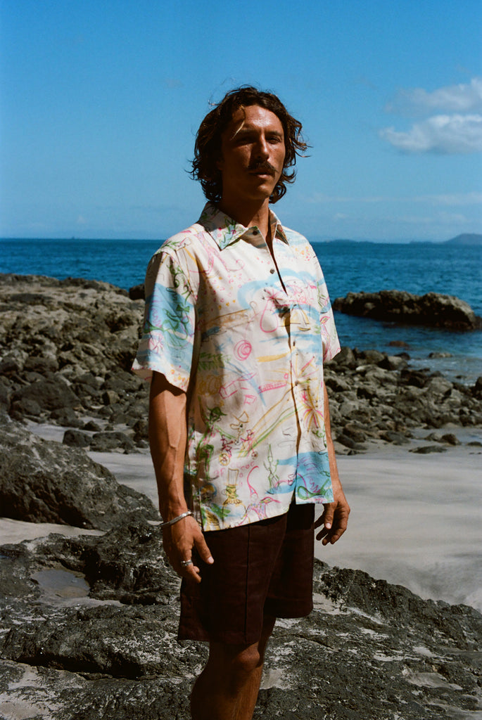 Cream-colored short-sleeve button-down shirt featuring an abstract print in pink, blue, yellow, and green, worn with dark brown shorts.