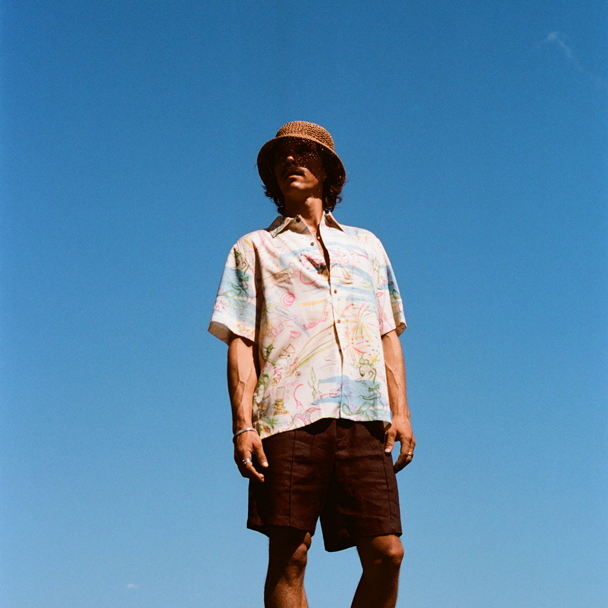 Man standing barefoot on rocks by the ocean, wearing a cream short-sleeve button-down shirt with a colorful abstract print and dark brown shorts. 