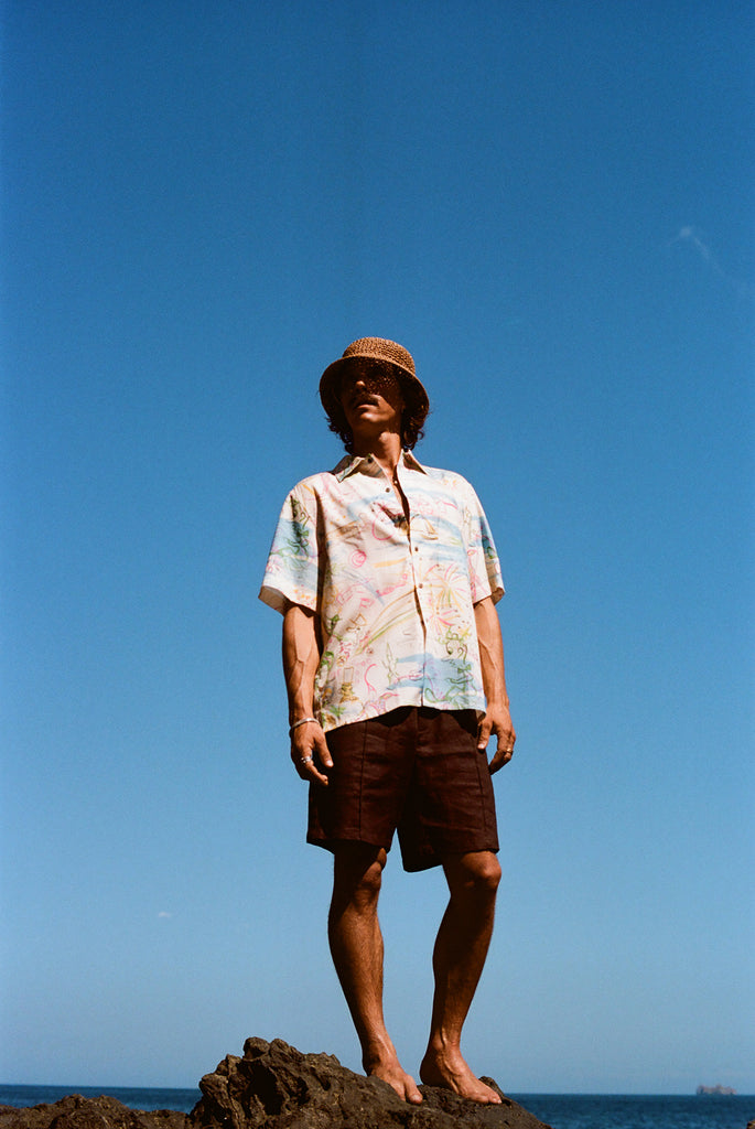 Man standing barefoot on rocks by the ocean, wearing a cream short-sleeve button-down shirt with a colorful abstract print and dark brown shorts. 