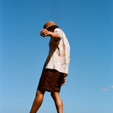 Man walking barefoot on black rocks near the ocean, wearing a cream button-down shirt with a colorful abstract print and dark brown shorts. 
