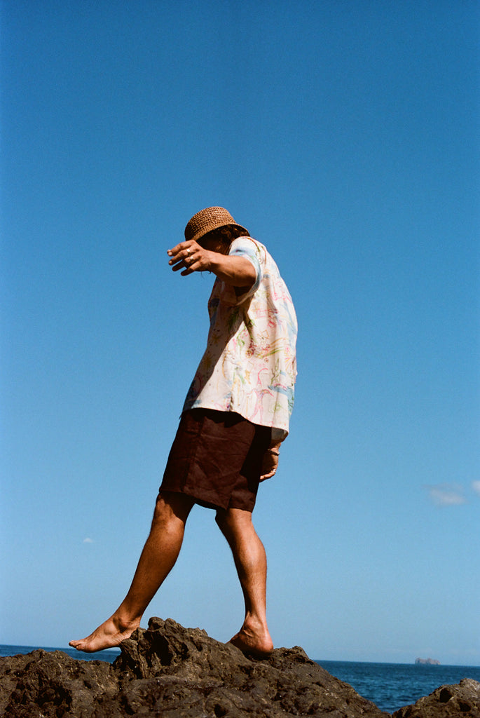 Man walking barefoot on black rocks near the ocean, wearing a cream button-down shirt with a colorful abstract print and dark brown shorts. 