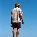 Man standing barefoot on black rocks with his back to the camera, wearing a cream button-down shirt featuring a colorful abstract print and dark brown shorts. 