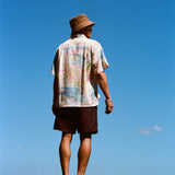 Man standing barefoot on black rocks with his back to the camera, wearing a cream button-down shirt featuring a colorful abstract print and dark brown shorts. 
