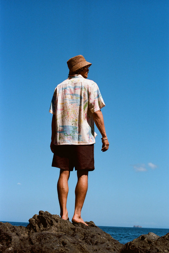 Man standing barefoot on black rocks with his back to the camera, wearing a cream button-down shirt featuring a colorful abstract print and dark brown shorts. 