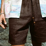Close up of male model wearing these brown linen shorts at the beach, wearing another Tombolo button-down open on top of the shorts.