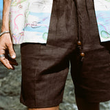 Close up of male model wearing these brown linen shorts at the beach, wearing another Tombolo button-down open on top of the shorts.