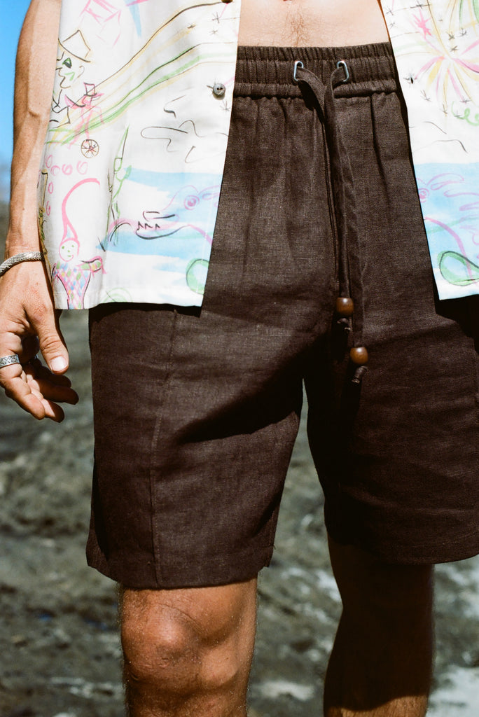 Close up of male model wearing these brown linen shorts at the beach, wearing another Tombolo button-down open on top of the shorts.