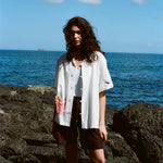 Woman standing on rocky shoreline wearing a white button-down shirt with embroidered fries in a red gingham pocket and fry details on the sleeves.